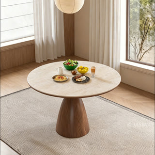 This elegant dining setting features a round stone-top table supported by a sturdy wooden pedestal base. The table is neatly set with bowls of salad, pasta, side dishes, and a drink. A textured area rug rests on the light wooden floor underneath the table, creating a warm and inviting atmosphere.