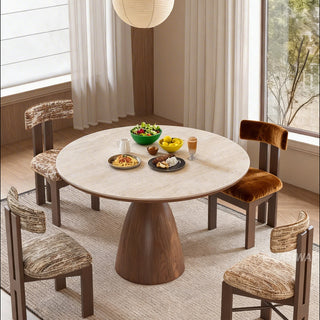 This elegant dining room features a round stone-top table with a sturdy wooden pedestal base. Four matching chairs with textured upholstery are arranged around the table, which is set with bowls of salad, pasta, and other dishes. A textured rug rests on the wooden floor beneath the furniture set, completing the warm and inviting space.