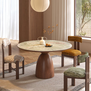 This modern dining room features a round green stone-top table with a sturdy wooden pedestal base. A white vase holding delicate branches and a small dish rest on the table surface. Four matching chairs with patterned upholstery are arranged around the table, set against a light-colored rug and wood-paneled accents.
