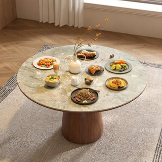 This round green stone-top dining table features a rich wooden pedestal base and rests on a textured area rug. The tabletop is abundantly laid out with multiple plates of food, bowls, a drink, and a central white vase holding slender branches. The surrounding room has light wooden herringbone flooring and white curtains, creating a warm, inviting dining space.