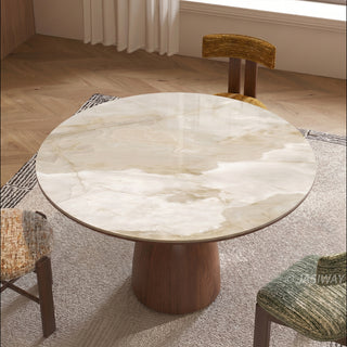 This overhead view highlights a round dining table featuring a polished stone top with natural marble patterns. The table rests on a sturdy, conical wooden pedestal base centered on a patterned area rug. Surrounding the table are matching chairs with upholstered seats in various textured fabrics.