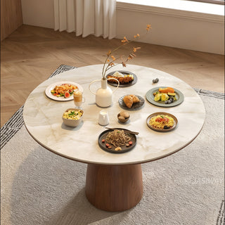 This round stone-top dining table features a rich wooden pedestal base and is placed on a textured area rug. The tabletop is abundantly laid out with multiple plates of food, bowls, a drink, and a central white vase holding slender branches. The surrounding room has light wooden herringbone flooring and white curtains, creating a warm, inviting dining space.