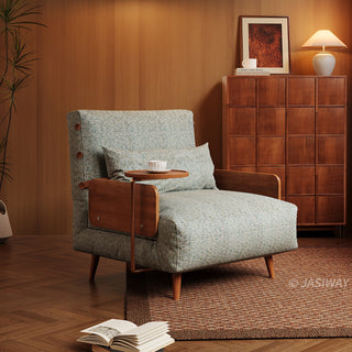 A modern armchair upholstered in textured sage green fabric sits on a patterned rug in a warm-toned room. The chair features wooden armrests, a matching lumbar pillow, and a small attached side table holding a teacup. In the background, a matching wooden cabinet topped with a lamp and framed artwork stands against wood-paneled walls.