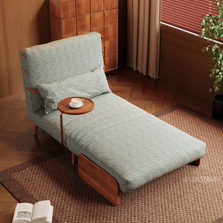 An elevated angle shows a modern sage green textured armchair extended into a chaise lounge configuration on a textured brown rug. The low-profile furniture features a matching lumbar pillow and an attached wooden side table holding a teacup. In the background, a tall wooden cabinet and a window with blinds complete the warm interior setting.