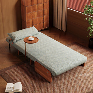 An elevated, high-angle view shows the modern sage green textured armchair fully reclined into a flat bed configuration. The low-profile piece rests on a textured brown rug, accompanied by a matching long bolster pillow and a small side table holding a teacup. In the background, a tall wooden cabinet and a window with blinds complete the warm, cohesive interior setting.