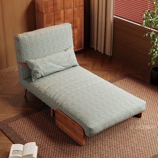 An elevated angle shows a modern sage green textured armchair extended into a chaise lounge configuration on a textured brown rug. The low-profile furniture features a matching rectangular lumbar pillow and dark wooden accents. In the background, a tall wooden cabinet and a window with blinds complete the warm, cohesive interior setting.