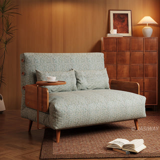 A modern loveseat sofa bed upholstered in textured sage green fabric sits on a patterned brown rug in a warm-toned room. The low-profile furniture features wooden armrests, two matching pillows, and an attached side table holding a teacup. In the background, a matching wooden cabinet topped with a lamp and framed artwork stands against wood-paneled walls.