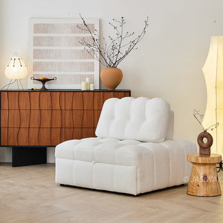 A white sherpa-upholstered modular armchair rests on a light wood floor in a modern living room. In the background, a warm wooden credenza holds abstract art, a decorative vase with branches, and a table lamp. A small wooden stump table stands beside the cozy seating piece.