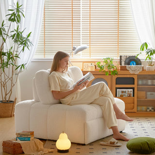 A woman sits comfortably on a white sherpa-upholstered modular armchair while reading a magazine in a bright, sunlit living room. The cozy seating rests on a patterned rug over a light wooden floor near a tall potted plant and a window with blinds. In the background, a wooden credenza displays decor, a record player, and books, creating a warm and inviting modern aesthetic.