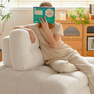A young woman relaxes comfortably on a white sherpa modular sofa, holding a green book in front of her face in a sunlit living room. The cozy tufted furniture rests near a light wooden floor with a wooden cabinet and potted plants in the background.
