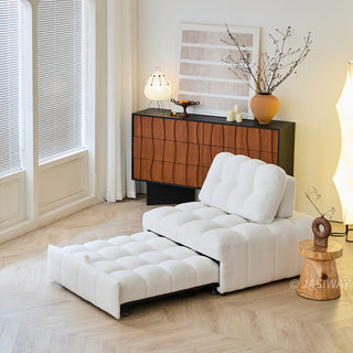 A white sherpa modular sleeper sofa bed is fully extended in a sunny, minimalist living room with a light wood floor. In the background, a warm-toned wooden credenza displays abstract wall art, a decorative vase with branches, and a table lamp.