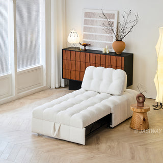 A convertible white sherpa lounge chair extends into a sleeper bed on a light wood floor in a brightly lit modern room. In the background, a two-tone wooden credenza holds abstract art, a decorative vase with branches, and a small lamp. A small wooden stump side table sits beside the furniture piece. The setting is warm and minimalist, showcasing functional, space-saving home decor.
