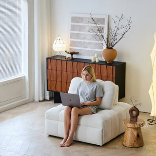 A woman works on a laptop while sitting comfortably on a white sherpa modular armchair in a brightly lit modern living room. The cozy seating arrangement rests on a light wood floor near a window with white blinds. In the background, a warm wooden credenza displays abstract wall art, a decorative vase with branches, and a small table lamp.