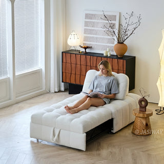 A woman sits comfortably reading a magazine on a white sherpa modular sleeper lounger in a sunlit modern living room. The convertible furniture piece extends across a light wood floor near a window with blinds. In the background, a warm-toned wooden credenza displays abstract wall art, a decorative vase with branches, and a table lamp.