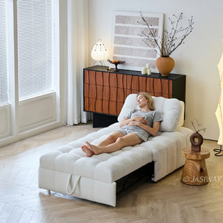 A young woman rests peacefully with her eyes closed on an extended white sherpa modular sleeper sofa bed in a sunlit modern living room. The cozy, tufted convertible furniture piece rests on a light wood floor near a window with blinds and a small wooden side table. In the background, a warm-toned wooden credenza displays abstract wall art, a decorative vase with branches, and a table lamp.