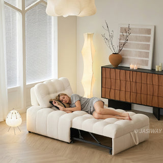 A young woman rests peacefully with her eyes closed on an extended white sherpa modular sleeper sofa bed in a sunlit modern living room. The cozy, tufted convertible furniture piece rests on a light wood floor near a window with blinds and a small wooden side table. In the background, a warm-toned wooden credenza displays abstract wall art, a decorative vase with branches, and a table lamp.