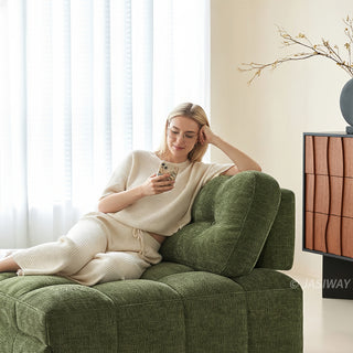 A young woman wearing glasses and a white knit outfit checks her smartphone while relaxing on an olive green tufted chenille convertible sleeper sofa in a bright modern living room.