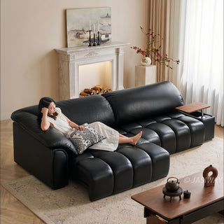A model relaxes on a modern black leather sectional sofa in a bright, warmly lit living room. The modular sofa features segmented cushioning, a built-in side table, and an extended chaise lounge section where she rests comfortably. The cozy interior background includes a fireplace mantel, a painting, and a wooden coffee table in the foreground.