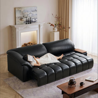 A model rests on a modern black leather sectional sofa in a bright, sunlit living room, with a book covering her face. The sofa features deep, segmented cushioning, an extended chaise section, and a built-in wooden side table. In the foreground, a wooden coffee table sits on a rug with a small tea set and a book. The background includes a fireplace mantel with a painting and a vase of cherry blossoms.
