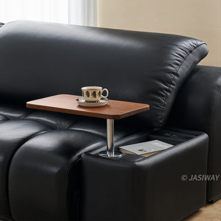 A close-up view shows a wooden swivel tray attached to the armrest of a modern black leather sofa. The rectangular tray table holds a patterned ceramic teacup and saucer. Beneath the armrest extension, a built-in storage compartment holds a magazine and control buttons.