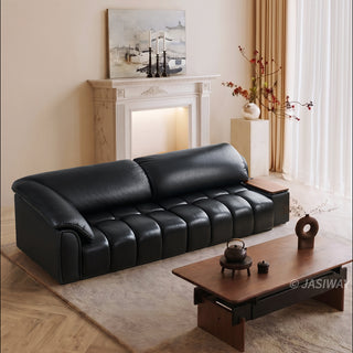 A modern black leather sofa is placed neatly in a cozy, warmly lit living room. The sofa features segmented cushioning and an integrated side tray. In the foreground, a wooden coffee table sits on a soft rug. The background includes a decorative fireplace mantel, a painting, and light-filtering curtains.