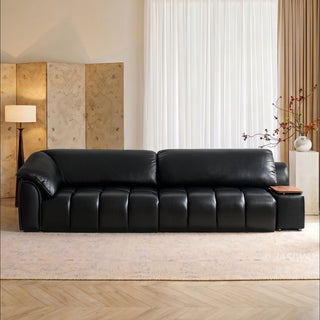 A modern black leather sofa is centered in a stylish, brightly lit living room. The sofa features distinctive segmented cushioning and an integrated wooden side table on the right armrest. It rests on a light-colored textured rug over a wooden floor. In the background, there is a decorative screen, a floor lamp, and a vase of cherry blossoms,