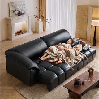 Two models are resting comfortably, lying side by side under a patterned blanket on a large black leather sectional sofa. The spacious modular sofa features deep, segmented cushioning and an extended chaise layout. In the foreground, a wooden coffee table sits on a textured rug. The warm, inviting living room background includes a fireplace mantel, a painting, and a floor lamp.