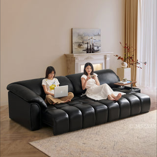 Two models are seated comfortably on a modern black leather sectional sofa in a well-lit living room. One woman sits on the left using a silver laptop, while the other sits on the right, holding a smartphone and relaxing. The sofa features distinct segmented cushioning and an integrated wooden side table holding a cup. The background includes a fireplace mantel with a painting, a floral vase, and sheer curtains.