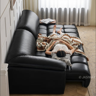 A high-angle view captures a model resting comfortably on a modern black leather sectional sofa. The sofa features plush segmented cushions and is partially covered by a patterned blanket with a pillow under the person's head. The cozy living room setting includes a textured rug on the floor and soft sheer curtains in the background.