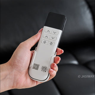 A hand with pink manicured nails holds a modern white and black remote control against a blurred black leather sofa background. The remote features six grey circular buttons arranged in three rows, controlling up and down movements.