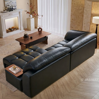 A high-angle view captures a modern black leather sectional sofa positioned in a bright, warmly lit living room. The modular sofa features distinctive segmented cushioning and an extended chaise lounge equipped with a built-in wooden side tray holding a cup. In the foreground, a wooden coffee table sits on a textured rug next to the sofa. The elegant background includes a fireplace mantel, sheer white curtains, and a floor lamp.