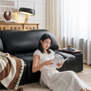 A model wearing a white dress sits comfortably on a textured rug, leaning against a modern black leather sofa while reading an open book. The plush sofa features segmented cushions and a built-in wooden side table holding a cup. The brightly lit living room background includes a wooden credenza, a unique modern table lamp, and soft sheer curtains.