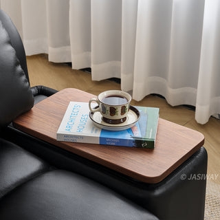 A close-up shot shows a built-in wooden side tray attached to a modern black leather sofa. Resting on the tray is a book titled "Architects' Houses" with a ceramic teacup and saucer placed on top of it. The background features soft, sheer white curtains filtering natural light into the room.