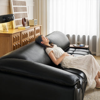 A model in a white dress relaxes comfortably with her eyes closed on a modern black leather sofa. The sofa features plush, segmented cushions and a built-in side tray holding a cup and a book. The brightly lit living room background includes a wooden credenza, decorative objects, and soft white curtains.