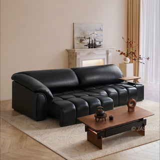 A modern black leather sofa is centered in a bright, warmly lit living room. The sofa features distinctive segmented cushioning and an integrated wooden side table on the right armrest. In the foreground, a low wooden coffee table sits on a textured rug, holding a small tea set and decorative items.
