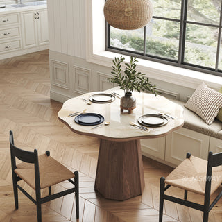 High-angle lifestyle shot of a modern round sintered stone dining table with a faceted wooden pedestal base, styled with place settings, woven chairs, and a window banquette in a warm kitchen and dining space.