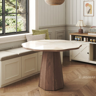 Lifestyle shot of a modern round sintered stone dining table with an organic-shaped top and a faceted wooden pedestal base, paired with a window banquette seating arrangement and herringbone flooring in a bright dining space.