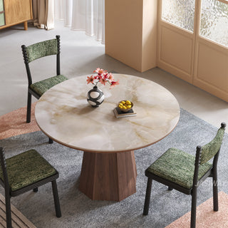 High-angle lifestyle shot of a round sintered stone dining table with a creamy onyx patterned top and a faceted wooden pedestal base, paired with green upholstered dining chairs and a patterned rug in a warm residential dining room.