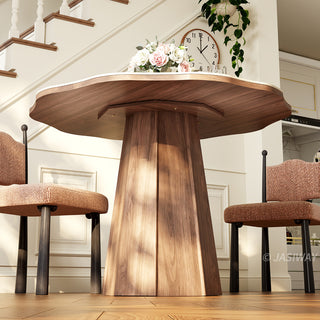 Low-angle detail shot of a modern round sintered stone dining table focusing on its organic-shaped tabletop, faceted wooden pedestal base, and textured upholstered dining chairs in a warm dining space.