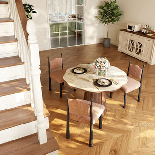 High-angle lifestyle shot of a modern round sintered stone dining table with a creamy onyx patterned top and a faceted wooden pedestal base, styled with textured upholstered chairs, place settings, and herringbone flooring near a staircase.