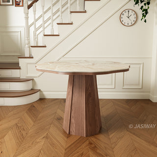 Front-facing product shot of a modern round sintered stone dining table with a creamy onyx patterned top and a faceted wooden pedestal base, placed on herringbone wood flooring in a residential interior.