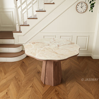 High-angle product shot of a modern round sintered stone dining table with a creamy onyx patterned top and a faceted wooden pedestal base, placed on herringbone wood flooring in a residential interior.